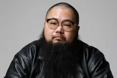 Um homem de cabeça raspada e barba longa preta, usando óculos de armação larga e jaqueta de couro preta, olha para a câmera