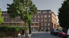 Vista externa de um prédio de cinco andares com ruas, carros e pedestres em primeiro plano