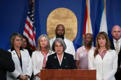 Pessoas em pé usando jalecos brancos. Uma mulher fala no microfone. Atrás há bandeiras dos Estados Unidos