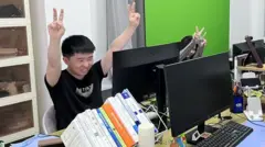 Shui Zhou, a young adult who pretends to have an office job, makes the victory sign with both hands that he is holding up in the air
