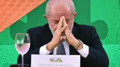Lula sentado, de olhos fechados, diante de fundo verde, apoia a testa nas mãos, encostadas como em um gesto de prece