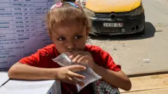 A girl holds a clear plastic bag filled with water as she sits atop a cart next to humanitarian aid packages provided by the United Nations Relief and Works Agency for Palestine Refugees (UNRWA) in central Gaza City on 27 August, 2024