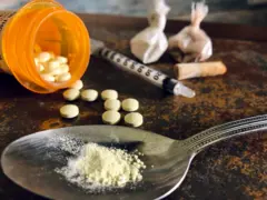 Frasco aberto de comprimidos, com alguns espalhados sobre a mesa, ao lado de uma colher com pó e uma seringa