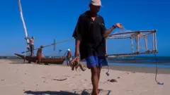 Pescador do litoral do Ceará caminhando na areia com algumas lagostas em uma mão e armadilha na outra