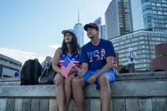 Una pareja observa desde un muelle el desfile de barcos Sail4th 250 en el puerto de Nueva York, con motivo del 250 aniversario de la Independencia de Estados Unidos, el 4 de julio de 2026. Ambos van vestidos con camisetas que llevan la bandera de Estados Unidos, llevan gorras. Uno de ellos lleva una bandera de Estados Unidos en la mano.