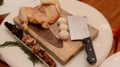 Faca de açougueiro com cabo preto ao lado de três cabeças de alho e meio frango cru sobre uma tábua de corte de madeira