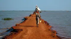 एक महिला अपने सिर पर भारी बोझ उठाए हुए पतली ज़ीरो-किलो 30 सड़क पर चल रही है, जो बेंटियू, यूनिटी स्टेट, दक्षिण सूडान के पास है