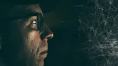 Um homem de óculos e de perfil tem parte de seu rosto iluminada enquanto o resto se funde com a escuridão ao fundo; ele encara um emaranhado de fios que se assemelha a uma teia de aranha.