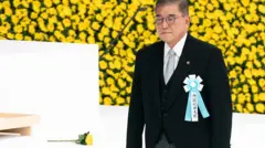 Исиба в черном сюртуке на фоне бело-желтых цветов.