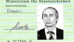 Um cartão de identidade da Stasi verde-claro traz a fotografia de Vladimir Putin em preto e branco à direita. Ele veste um terno escuro e uma gravata listrada, tem cabelos loiros e olha para o horizonte, fora do enquadramento.