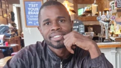 Yostin Mosquera repousa sua mão esquerda contra o queixo e sorri para a câmera, sentado dentro de um bar com mesas e cadeiras