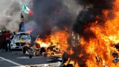 Manifestante no alto de um carro exibe uma bandeira do México, enquanto veículos são incendiados em uma rua de Los Angeles, em 8/6/2025