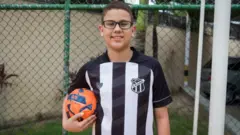 Um menino usando óculos de grau e uma camisa de futebol do Ceará. Ele segura uma bola de futebol com a mão direita.