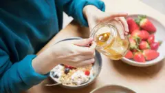 Una persona colocando miel sobre un plato de yogur, cereales y frutas