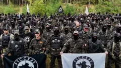 Dezenas de homens usando roupas militares e segurando bandeiras pretas e brancas com a imagem de um cavaleiro montado posam diante da câmera, com uma área de floresta ao fundo
