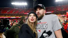 Taylor Swift i Travis Kelce podczas meczu o mistrzostwo AFC na stadionie GEHA Field w Arrowhead Stadium w Kansas City w stanie Missouri. Swift ma długie, jasne włosy, ciemną bluzę i ciemną czapkę na głowie, Kelce ma szarą koszulkę i ciemną czapkę z daszkiem.