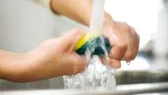 Foto de uma pessoa torcendo uma esponja de cozinha debaixo da torneira.