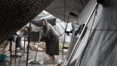 Una mujer descalza intenta barrer el agua que inunda las carpas