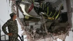A man in camouflage uniform and hat looks at the wreckage of the plane. He is standing in front of a wall with a gaping hole