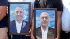 Двое мужчин держат два портрета в коричневых рамках. На левом портрете изображен бритый мужчина в синем пиджаке и белой рубашке. На правом портрете мужчина в черном пиджаке и белой рубашке