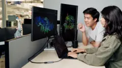 Duas pessoas trabalhando juntas em uma mesa, em um ambiente que parece ser de laboratório ou escritório de pesquisa. Grandes monitores de computador exibem modelos coloridos de estruturas moleculares ou proteicas tridimensionais sobre fundos escuros, enquanto um laptop sobre a mesa mostra linhas de código ou dados. Uma pessoa está sentada usando o laptop, e a outra está inclinada em direção às telas, ambas concentradas nas visualizações científicas. Prateleiras e equipamentos de laboratório são visíveis ao fundo, sugerindo um ambiente de pesquisa biomédica, biotecnológica ou computacional. A cena enfatiza o uso de ferramentas de análise e visualização baseadas em computador para o estudo de estruturas biológicas complexas.