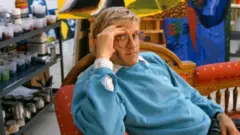 David Hockney 1987'de Kaliforniya'daki Stüdyosunda mavi bir kazak giymiş, eli yüzüne bakacak şekilde sandalyede dinleniyor.
