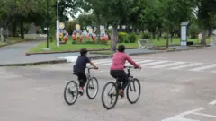 Duas crianças andam de bicicleta em uma rua larga, em frente ao monumento com bonecas do tipo matrioskas ao fundo