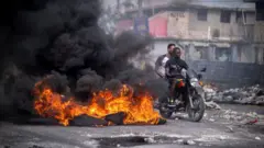Dos personas en moto en puerto príncipe transitan entre basura y una pila de neumáticos encendidos 