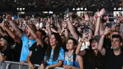 Fãs do Oasis no Principality Stadium. Um grande número de pessoas pode ser visto olhando para o palco. Muitos têm as mãos no ar e estão gravando o show em seus smartphones.