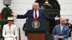 Donald Trump stoi przy mównicy prezydenta Stanów Zjednoczonych, mając uniesione obie ręce na boki. Po jednej stronie siedzi król Karol, po drugiej żona Trumpa, Melania. Król ma na sobie jasnoniebieski garnitur w delikatne paski, jasną koszulę oraz jasnoniebieski krawat z pasującą poszetką; patrzy w bok i śmieje się. Trump ubrany jest w granatową marynarkę, białą koszulę i czerwoną krawatkę, przemawiając do zgromadzonych. Melania ma na sobie dopasowany biały garnitur oraz słomkowy kapelusz. Scena rozgrywa się przed Białym Domem.