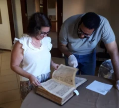 Pesquisadores analisam um manuscrito antigo em uma mesa de trabalho. Ela folheia as páginas enquanto ele observa atentamente, usando luvas de proteção para manusear o documento histórico.