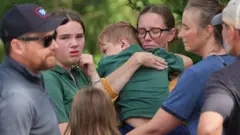 Несколько взрослых и несколько детей, в центре кадра женщина, которая прижимает к себе мальчика, пытаясь его успокоить