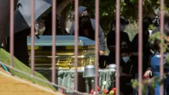 Золотой гроб и люди в масках на похоронах Эль Менчо. Фото сделано сквозь решетку ограды