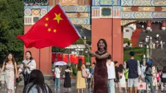 2025年10月7日國慶黃金周假期期間，一位女士在重慶人民大禮堂前揮舞著一面巨大的中國國旗，訪客們匯聚於這一地標建築。廣場上遍布愛國裝飾和紅旗，人們共同慶祝中華人民共和國成立76週年。