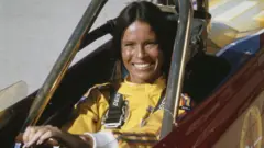 基蒂·奧尼爾（Kitty O'Neil）——一位留著烏黑直髮的女性——坐在一輛敞篷車中，身穿黃色連身衣，雙手握著方向盤，對著鏡頭露出微笑（7/7/1977）