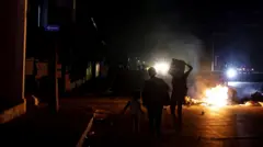 Três crianças estão diante de uma pilha de lixo em chamas no meio de uma rua em Havana. A rua está completamente às escuras, exceto pelos faróis distantes dos carros.