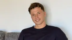Freddie Woodward: un joven de pelo corto y oscuro está sentado en un sofá gris, sonriendo a la cámara. Tiene el pelo corto castaño y lleva un jersey azul marino.