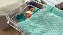 Um bebê dorme em uma cama de hospital em Kiev, usando um gorro amarelo e coberto por um cobertor verde-claro