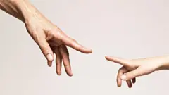 Mão de pessoa mais velha e de criança quase se tocando como na pintura de Michelangelo
