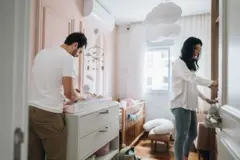 Pai trocando fralda em cômoda, enquanto mãe mexe em armário