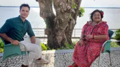 Dona Onete com vestido vermelho estampado se senta à frente do repórter João Fellet, que veste camisa verde e calça clara; ao fundo deles, vê-se o tronco de uma árvore e um rio bastante largo