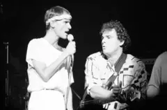 Cazuza cantando no microfone ao lado de baixista, que parece cantar também