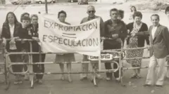 Un grupo de personas, principalmente mayores, están de pie junto a una baranda que parece cerrar el acceso a una calle. En el centro dos de ellos cargan una pancarta que dice "expropiación=especulación".