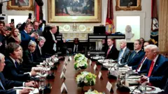 Milei e Trump, entre outros homens, sentado frente a frente em mesa durante reunião