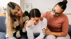 Duas jovens confortam a amiga que está sentada entre elas no sofá, com a mão no rosto e expressão abatida