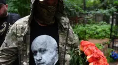 مردی با چهره پوشیده با اورکت نظامی و پیراهنی که تصویر پریگوژین روی آن هست