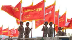 Soldados em pé, em uniforme de combate, na parte traseira de caminhões, segurando grandes bandeiras vermelhas com dizeres em chinês
