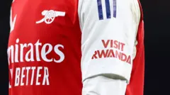 Un maillot rouge et blanc d'Arsenal est visible avec un logo Visit Rwanda sur la manche gauche.