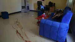 A long smear of blood spreads across a stone floor. A blue sofa, a table with some cups on it and an office chair and table can also be seen around the outside edge of the room