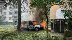 У дворі житлового будинку горить білий фургон: з-під капота і салону виривається вогонь і густий дим. Навколо — повалене листя й уламки, поруч стоять дерева, а на тлі видно багатоповерхівку з пошкодженими вікнами.